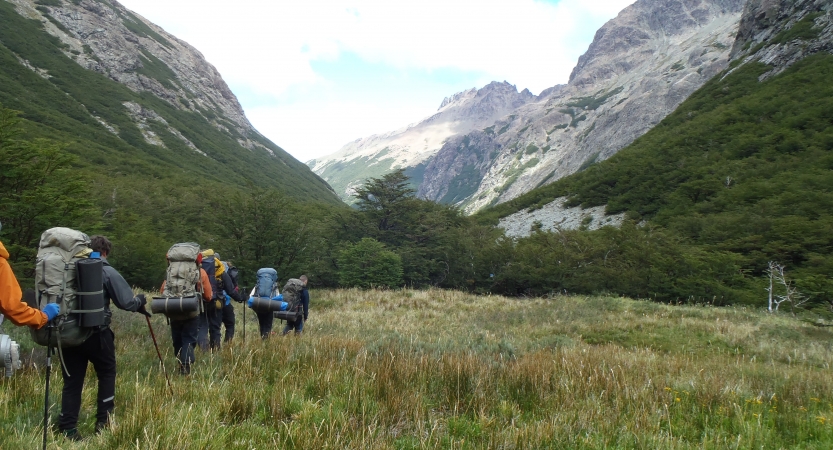 A group of people wearing backpacks hike through a meadow, away from the camera. Tall mountains frame the meadow. 